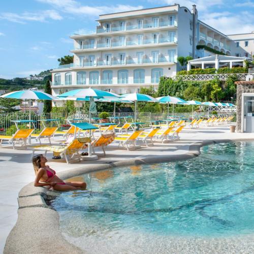 Piscina all'aperto con lettini e ombrelloni, hotel sullo sfondo, giornata soleggiata.