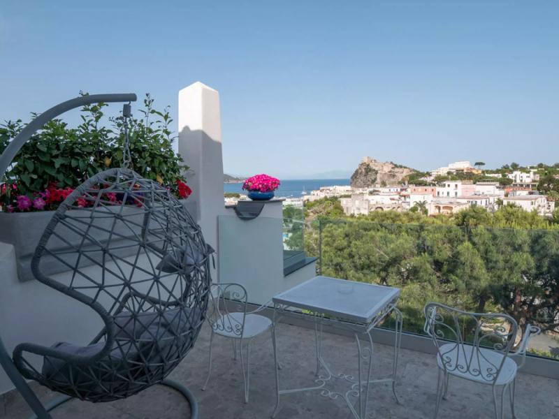 Terrazza con vista mare, sedia sospesa e tavolino, fiori colorati e panorama.