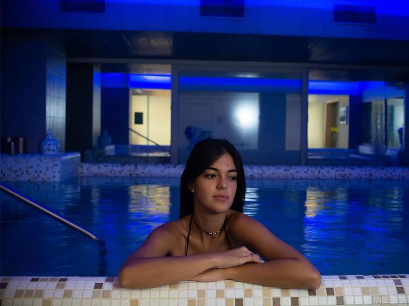Ragazza in piscina illuminata di blu, appoggiata al bordo e pensierosa.